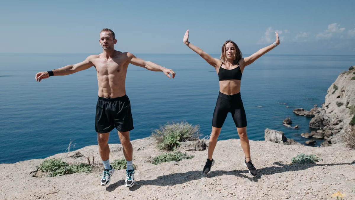 A man and woman perform jumping jacks on a cliff by the ocean under a clear sky.